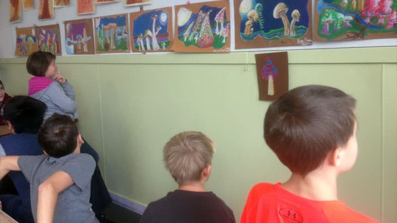 Children in art classroom lined up and looking at drawing of mushrooms