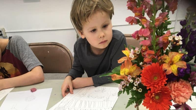 A lightskinned boy around age 5 with blond hair leaning over a pice of paper. He has a pencil in is hand and is looking intently at an arrangement of red and yellow flowers.