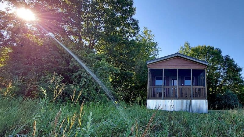 small wooden cabin with screen-in porch on grassy hill with bright sun and blue sky