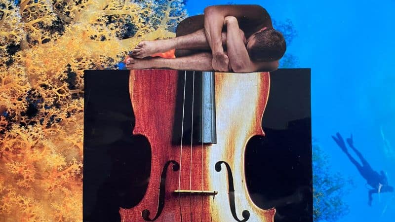 Naked Man curled up on top of image of violin on unwater background mainly blues and yellow colors