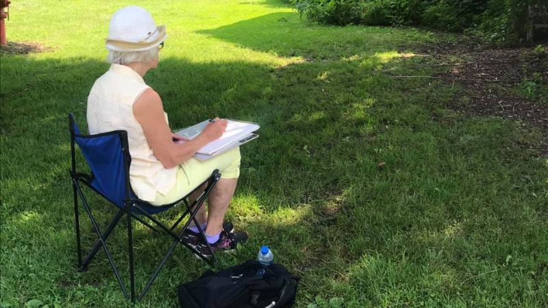 Older white woman dressed in white sitting in a camping chair drawing