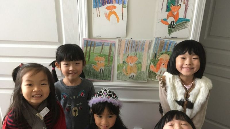 Five young asian girls smiling. They are standing infront of a wall with their watercolor paintings of foxes. One girl is wearing a crown. 