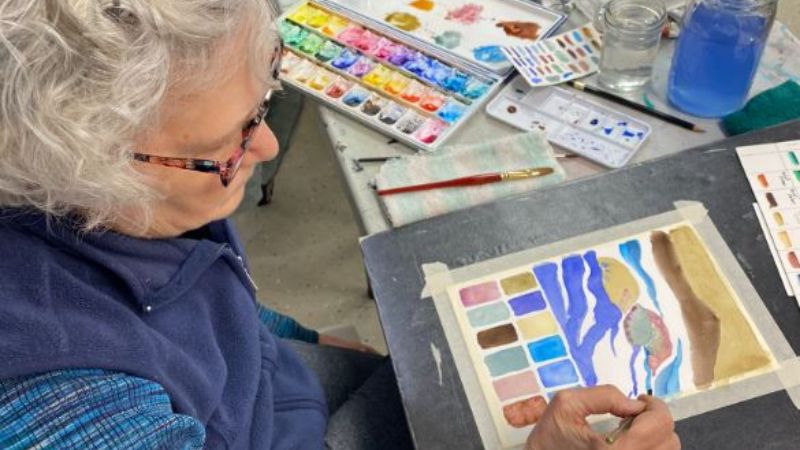 Woman with grey hair dressed in blue painting with a colorful abstract watercolors. There are pan pallets and water on the table.