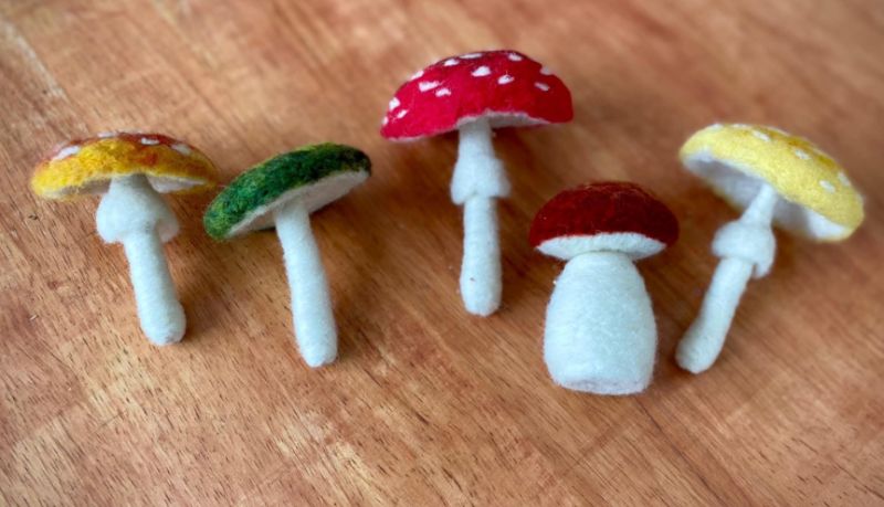 five 3-D felt mushroom in a row on a wood floor. The stems are white the tops are yellow blue and red with white spots.